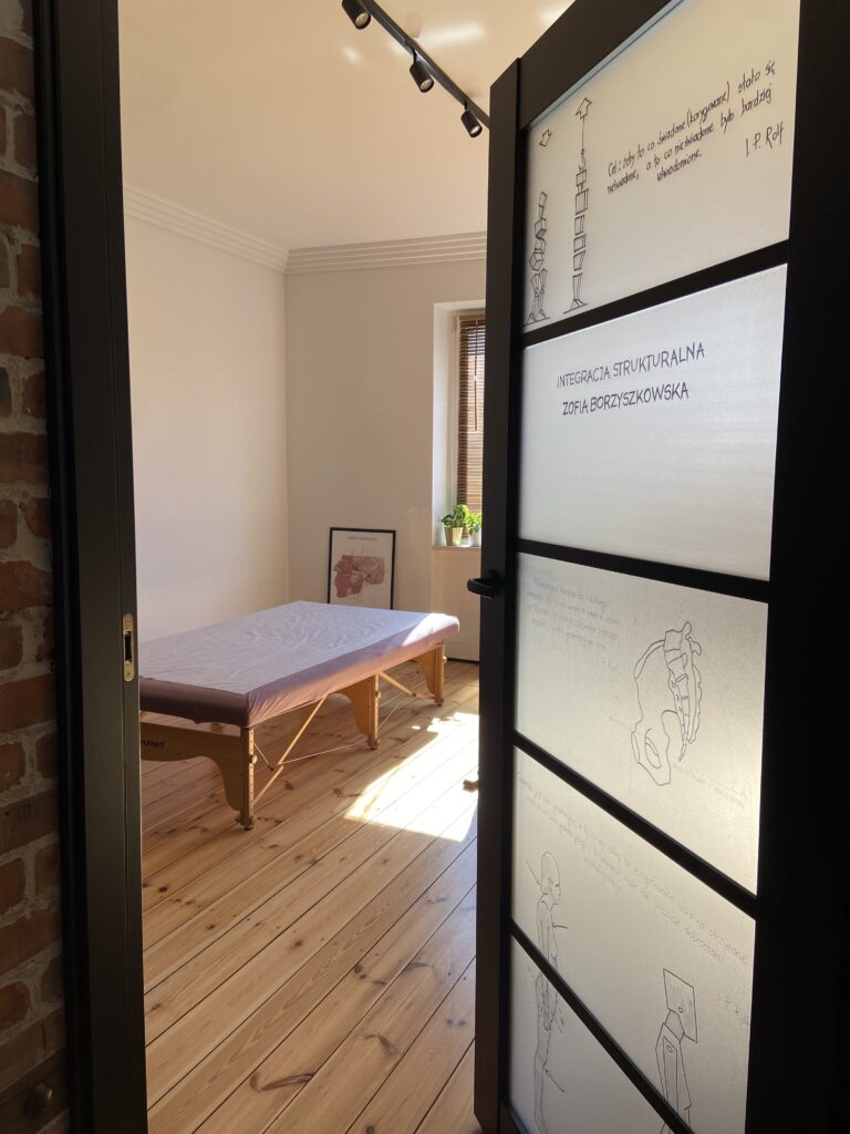 Wnętrze jasnego gabinetu terapeutycznego z profesjonalnym stołem do masażu i szklanymi drzwiami z grafikami anatomicznymi